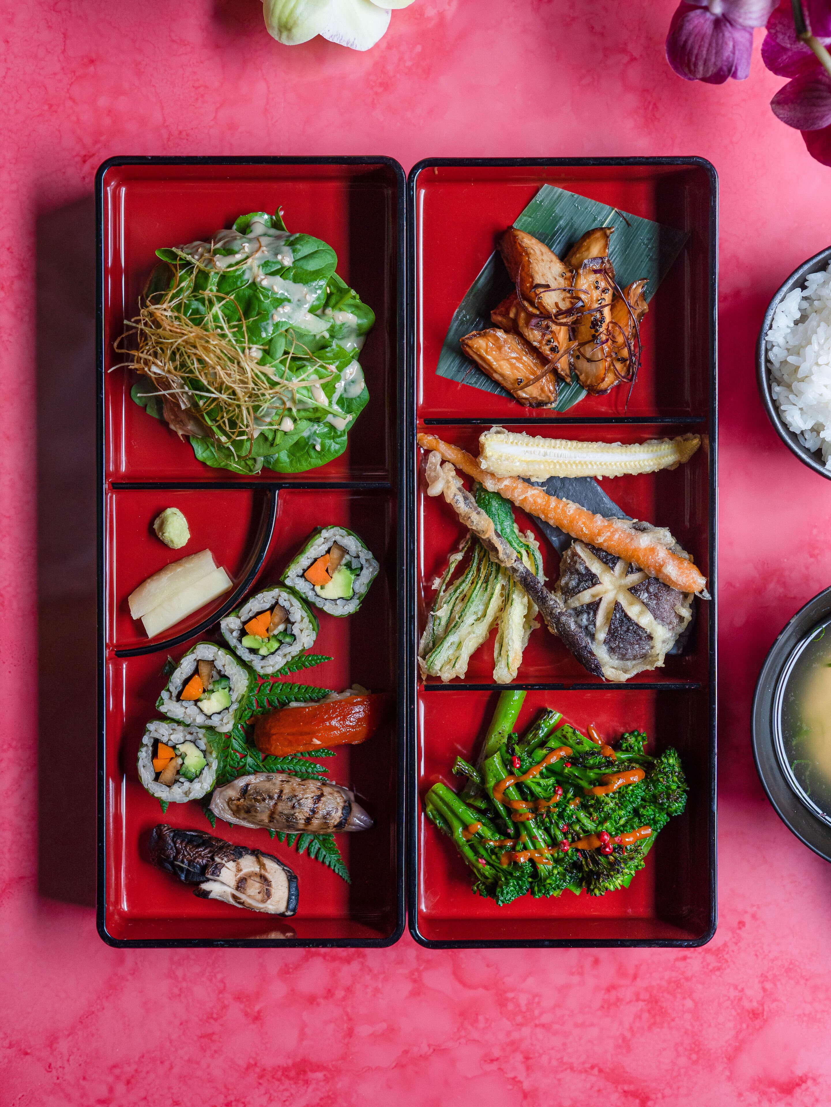 A bento box with various sushi and vegetables on a red tray.