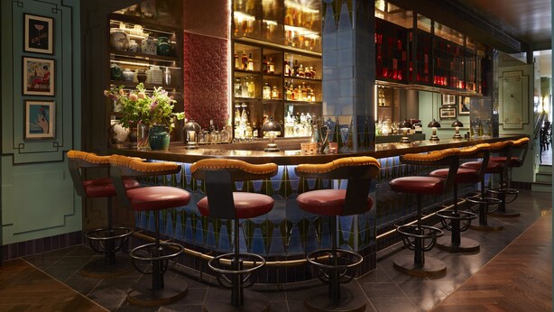 A bar area with multiple stools, a bar counter, and shelves stocked with bottles. - Beaverbrook Town House (London)