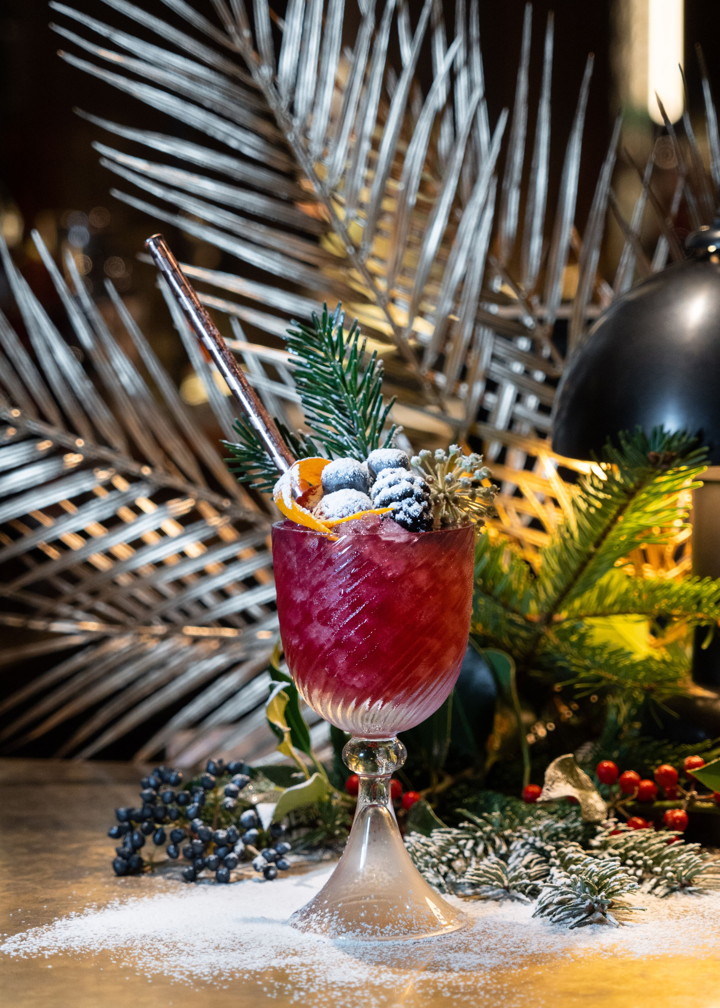 A festive drink garnished with pine branches, berries, and an orange slice, set against a backdrop of pine branches and a Christmas ornament.