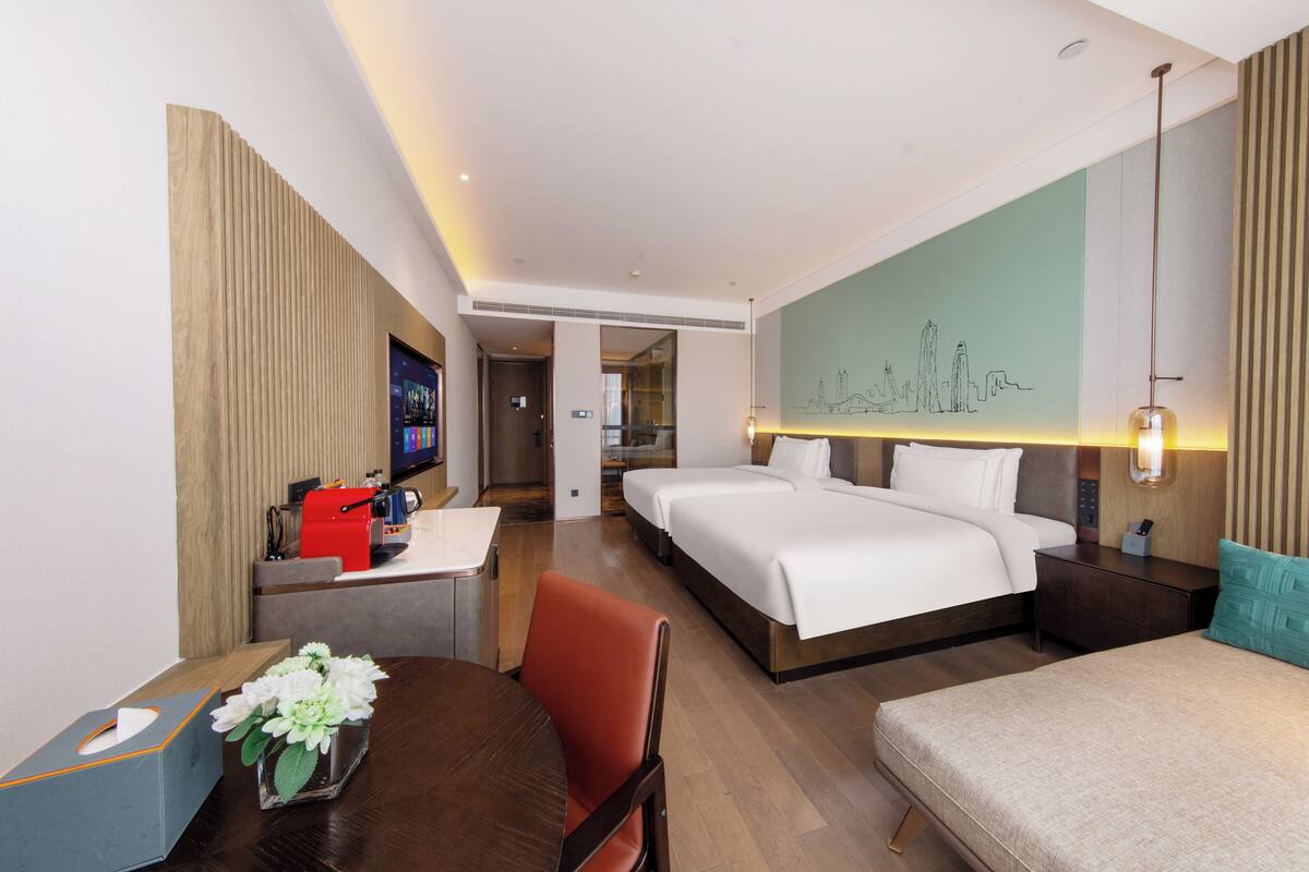 A hotel room with a bed, bedside tables, a desk with a coffee machine, a chair, and a decorative vase.