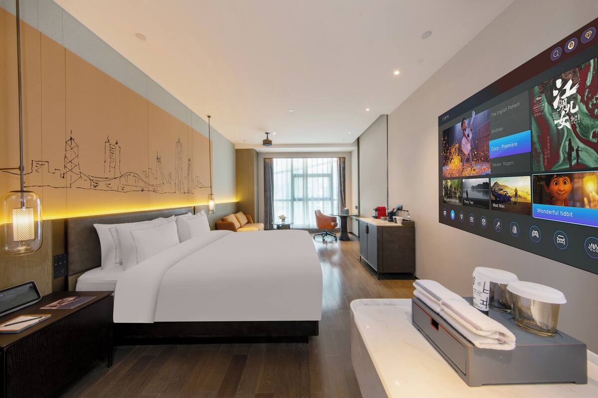 A modern hotel room with a large bed, a desk with a laptop, a TV displaying a movie, and a wall-mounted TV.