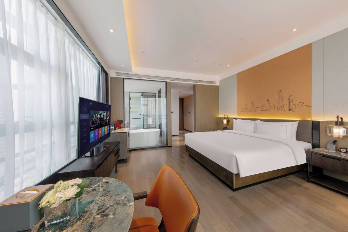 A modern hotel room with a large bed, a desk with a computer, a television, and a bathroom visible through an open door.