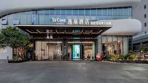 Una entrada de hotel moderna con un amplio frente de vidrio, señalización de 'UrCove by Hyatt' y un área exterior bien cuidada con plantas y una estatua decorativa.
