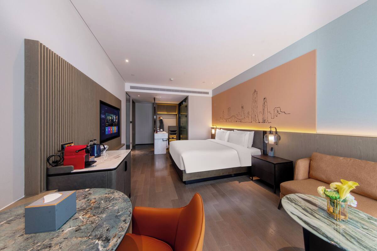 A modern hotel room with a bed, a desk, a sofa, and a wall-mounted TV.