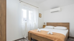 A bedroom with a wooden bed, white bedding, a white air conditioner, a bedside lamp, and a window with sheer curtains. - Centrally-located two-bedroom apartment w/sunny terrace Marina IV. (Split)