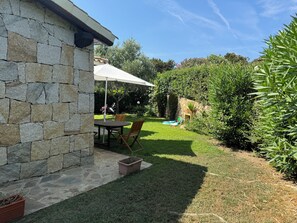 Eine Steinterrasse mit Tisch und Stühlen, einem überdachten Sonnenschirm und einem kleinen Garten mit Rasen und verschiedenen Pflanzen.