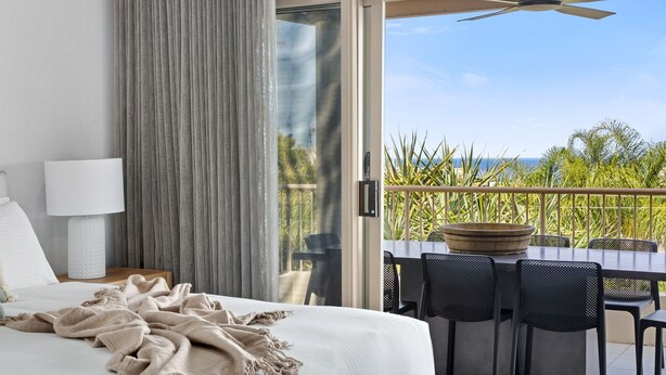 A hotel room with a bed, a nightstand, a lamp, a balcony with a table and chairs, and a view of the ocean.