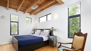 Un dormitorio con cama, mesitas de noche, una silla y ventanas amplias.