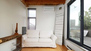 Un sofá blanco en una esquina, vigas de madera en el techo, una ventana con vistas y una pared blanca con un equipo de aire acondicionado.