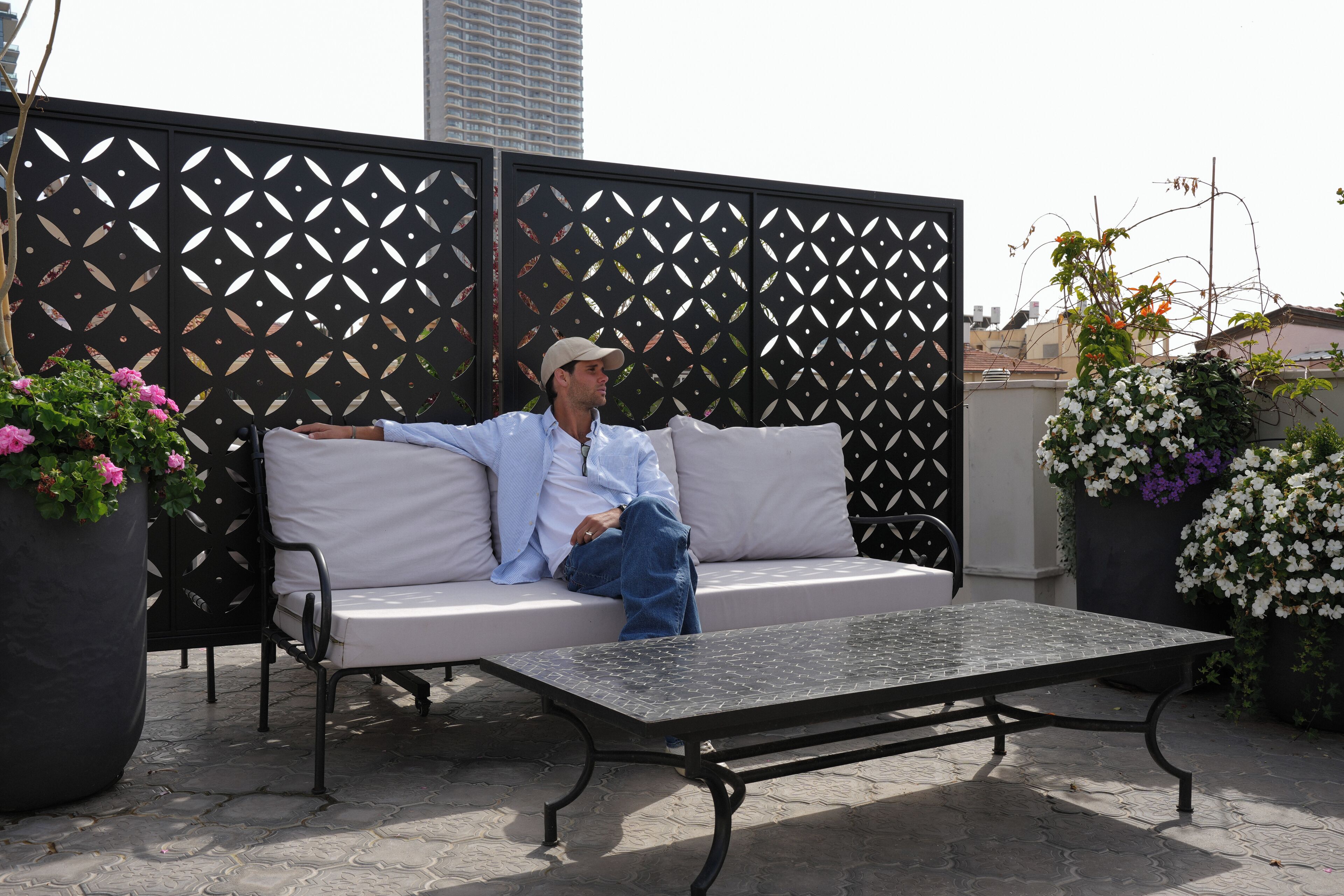 A man sitting on a white sofa with a metal table in front of a decorative black fence.