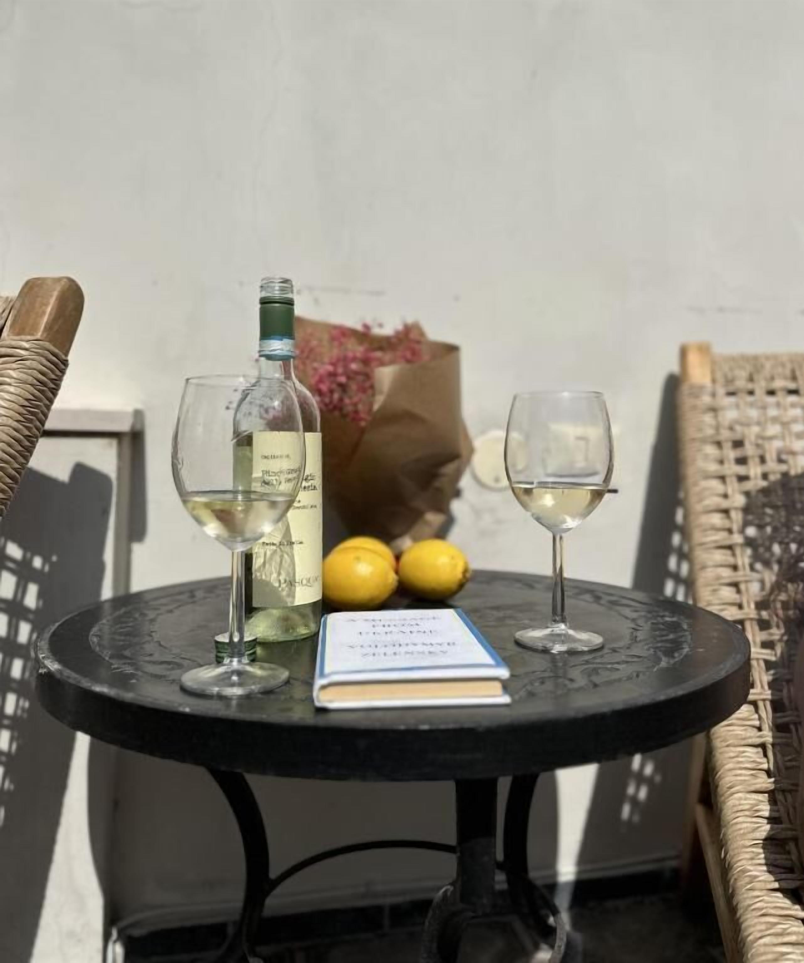 A small round table with two wine glasses, a bottle of white wine, and two lemons. A book is also on the table.
