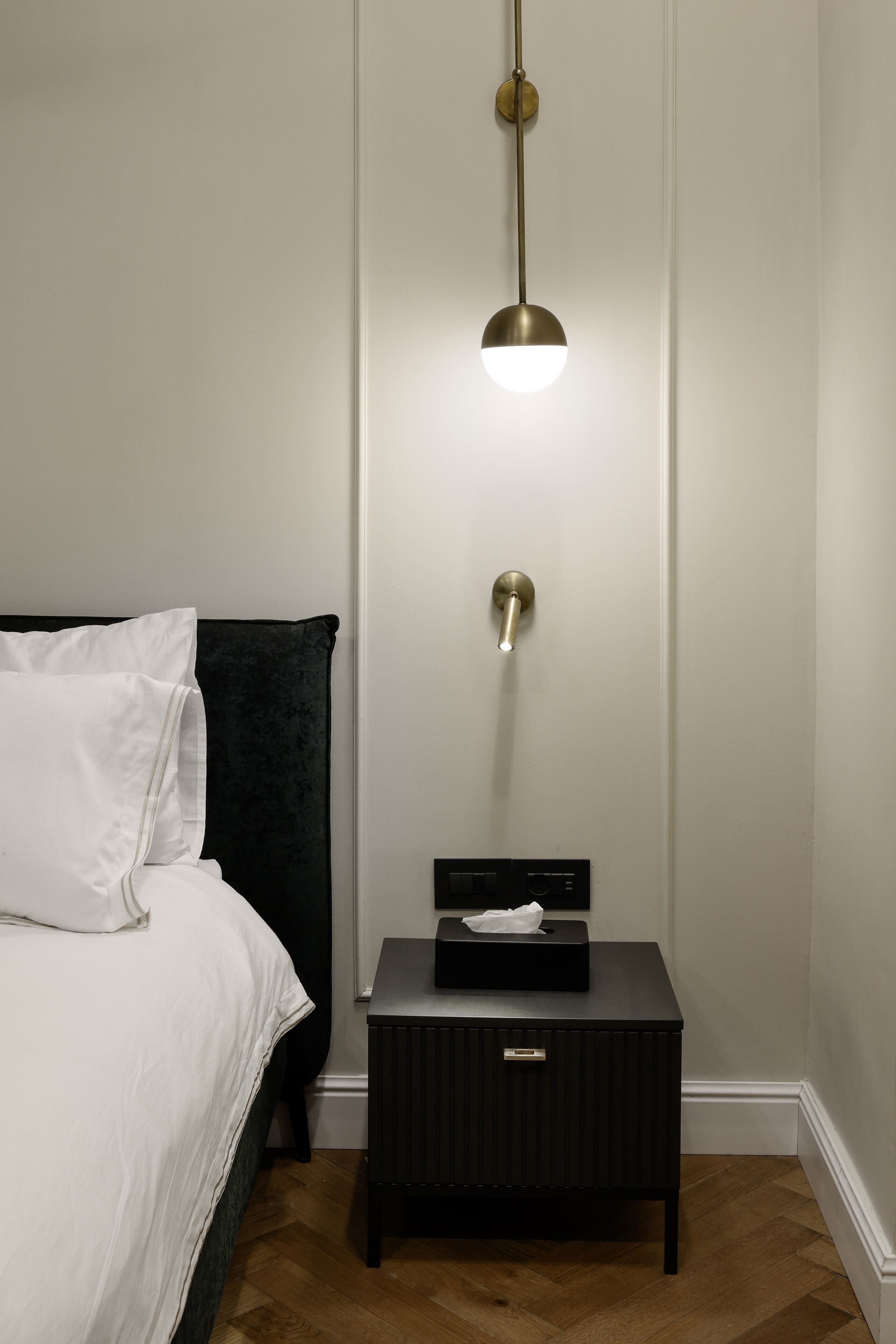 A neatly made bed with white linens, a black headboard, a bedside table with a tissue box, and a wall-mounted light fixture.