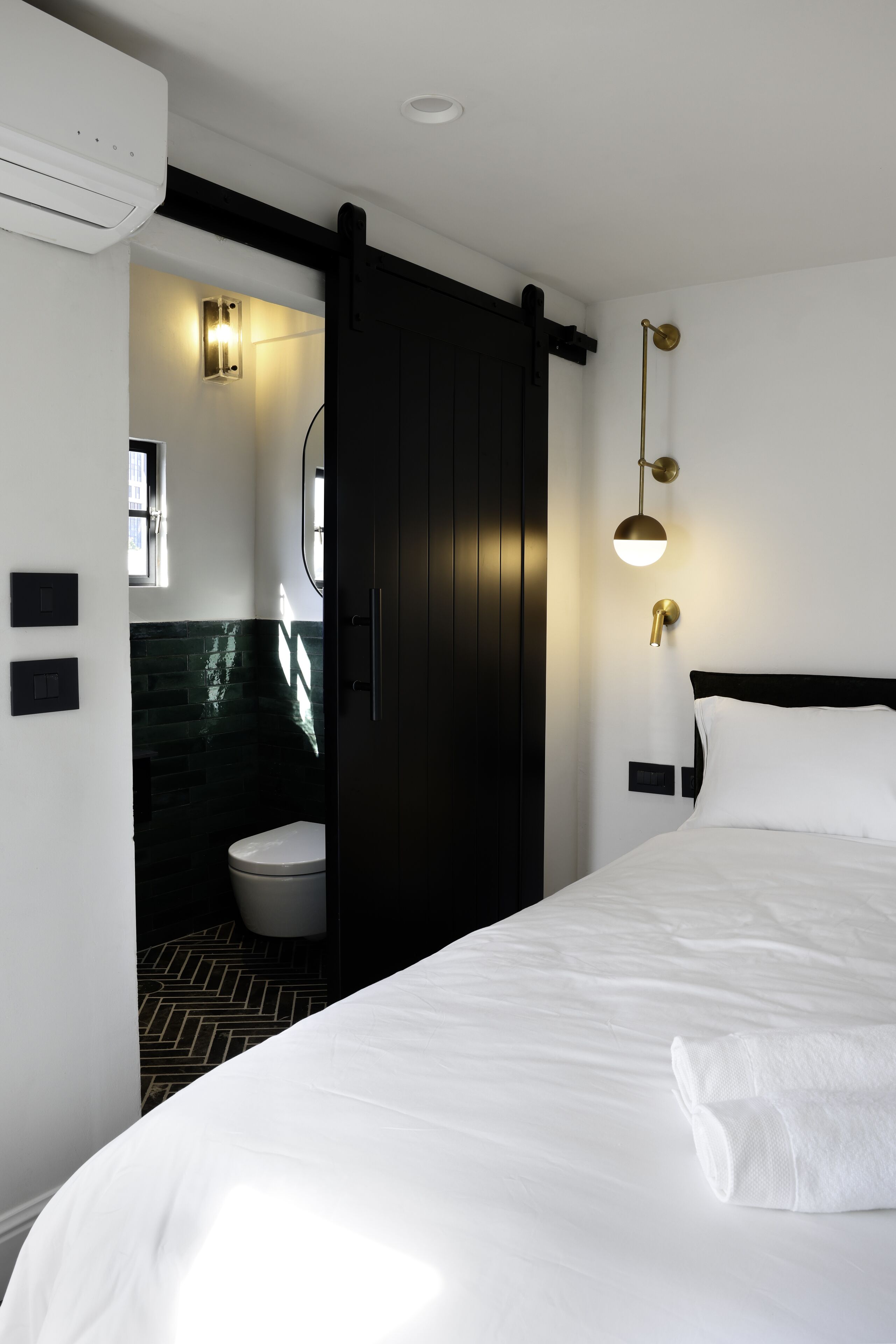 Una habitación de hotel moderna con una cama grande, una puerta negra que da al baño y una lámpara fijada a la pared.