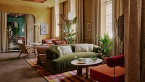 A spacious living room with a green sofa, red armchair, and a small table with a glass. There are large windows with curtains, a ceiling with wooden panels, and a potted plant. - Ember Locke Kensington (London)
