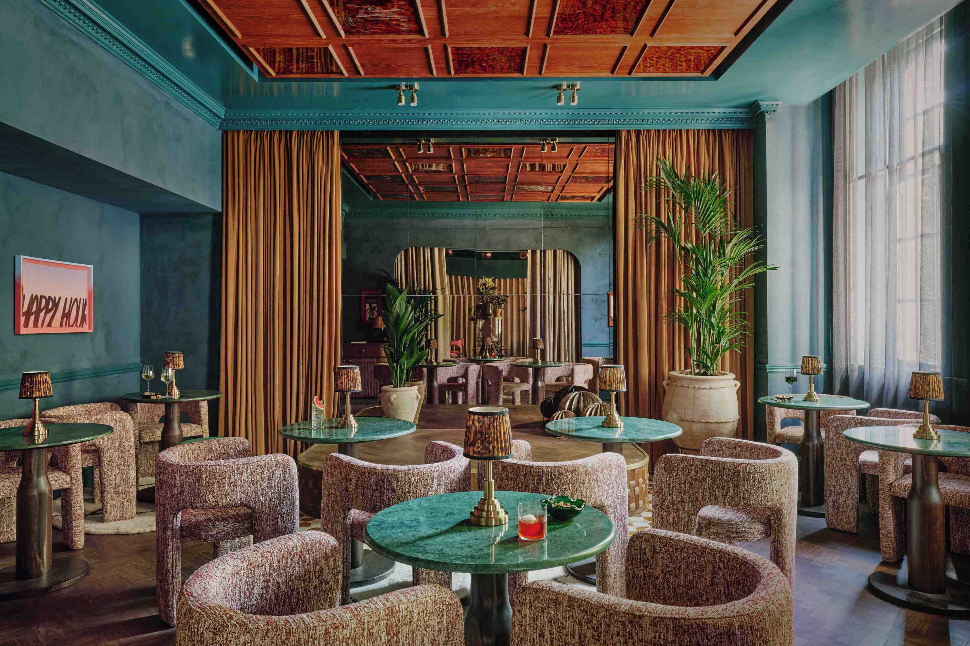 A spacious lounge area with round tables and upholstered chairs, large potted plants, and a view into the dining area.