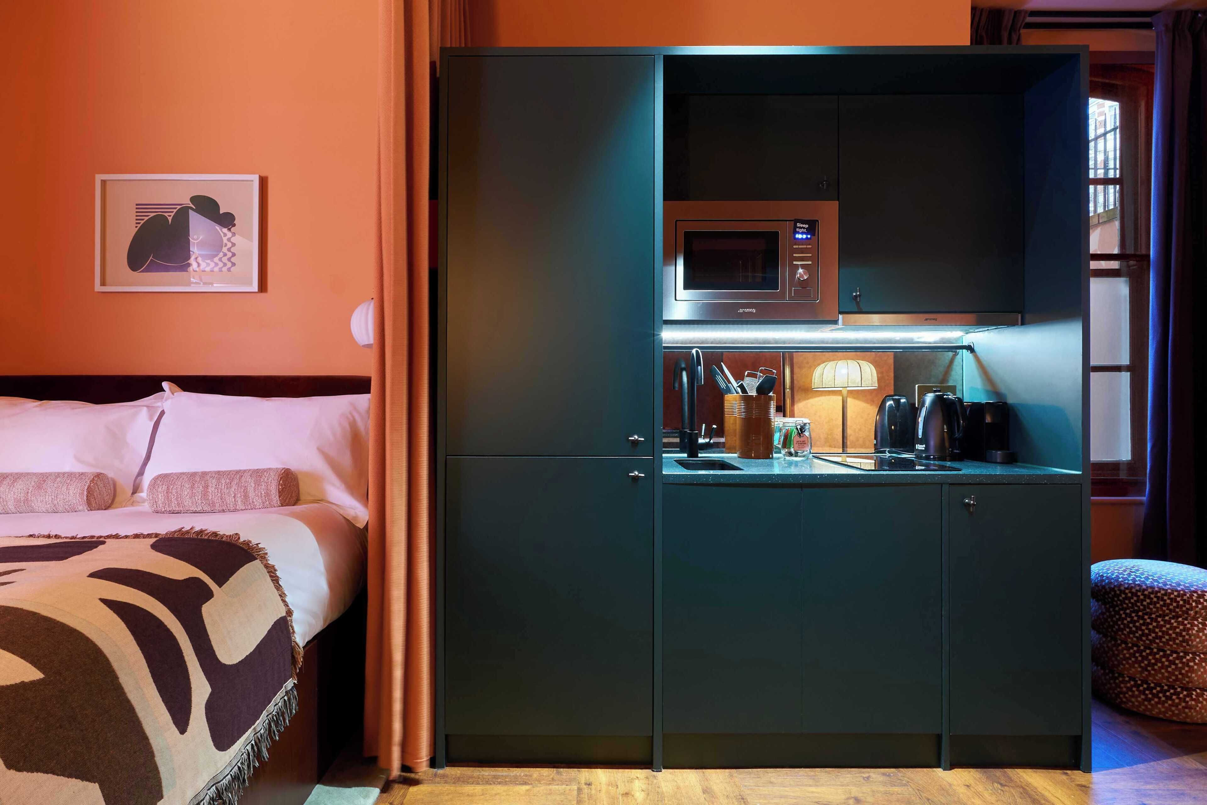 A hotel room with a bed, a kitchenette with a microwave, a coffee maker, and a small table with a lamp.