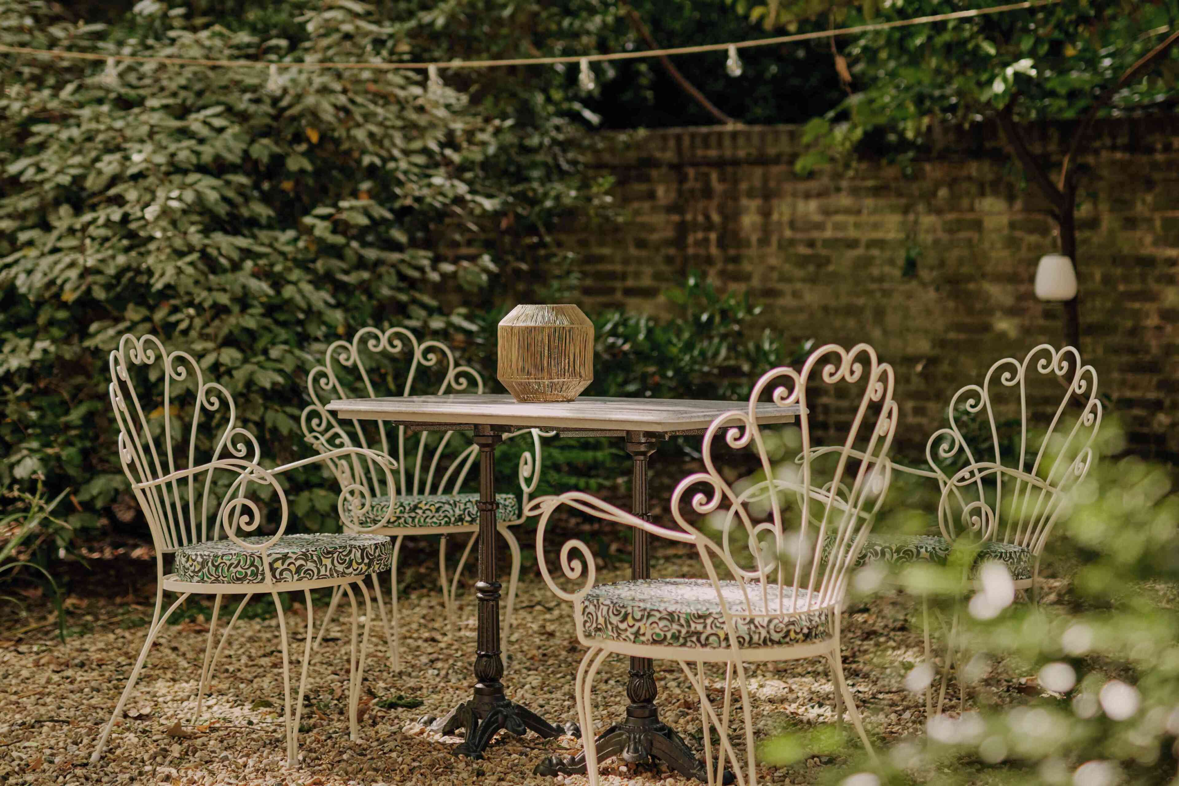 A garden patio with a table and four chairs, surrounded by greenery and a brick wall.
