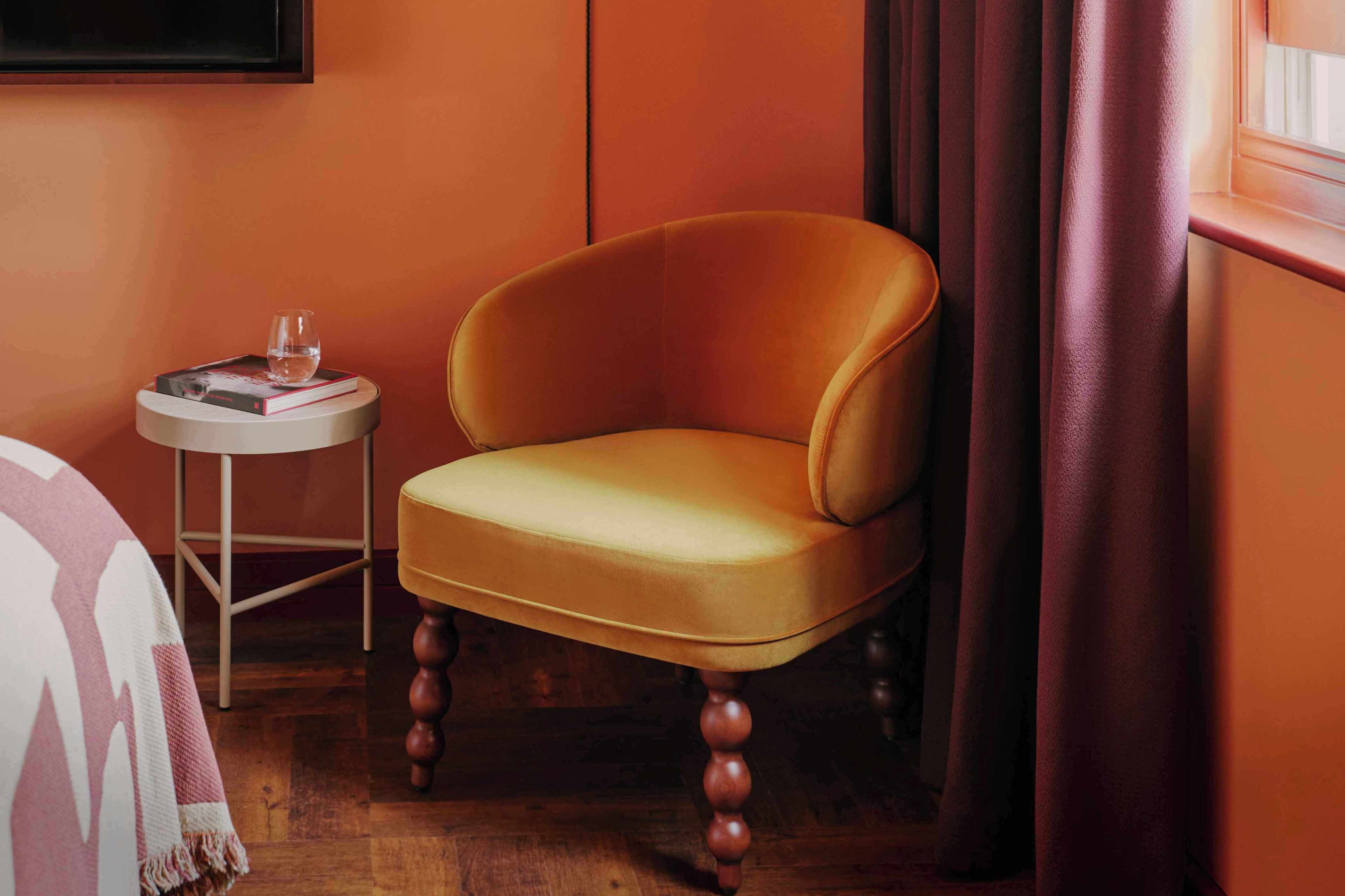 A room with a single chair, a small round table with a glass and book, and a window with curtains.