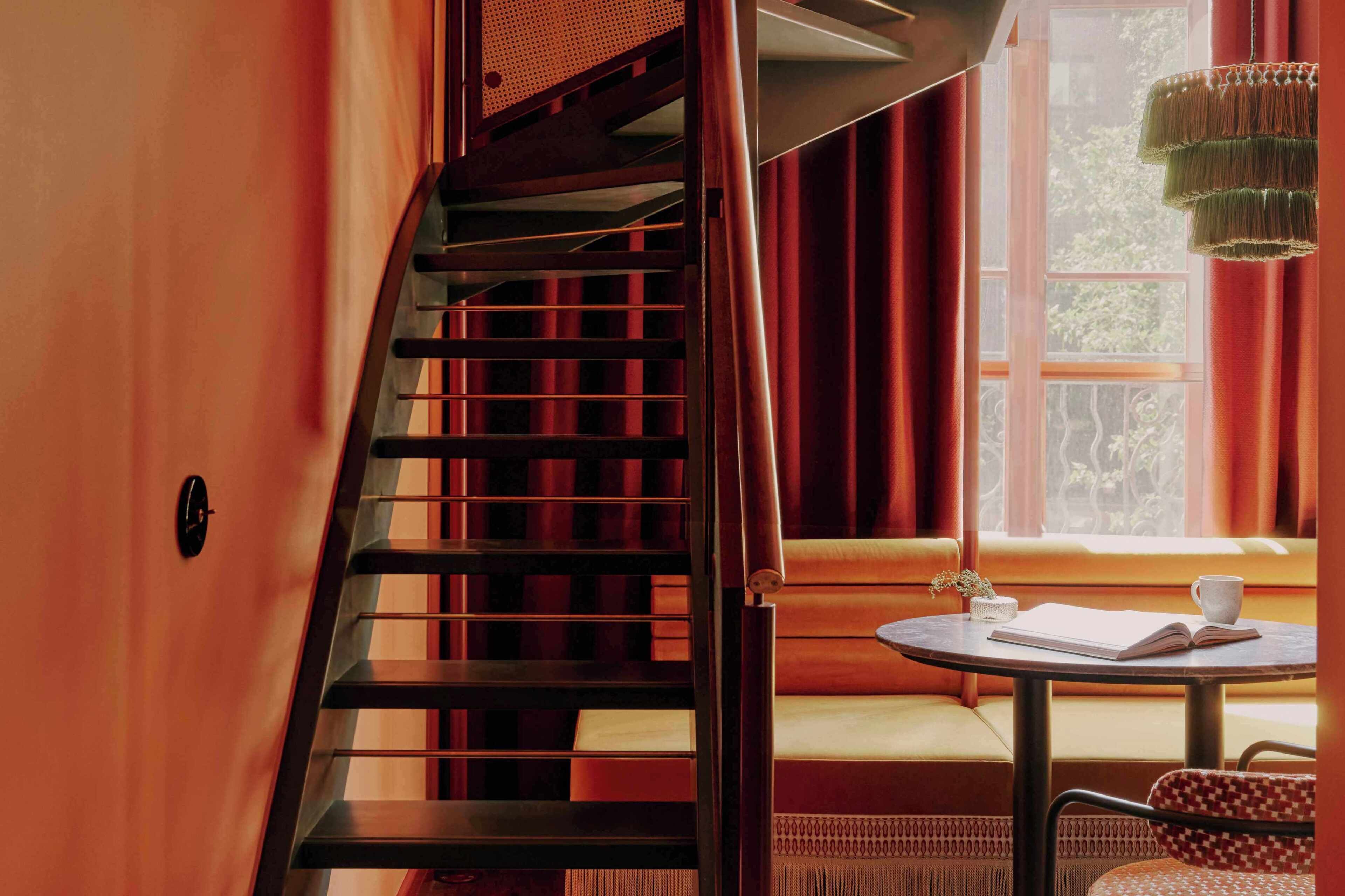 A staircase, a window with curtains, a table with a book and a cup, and a chair.