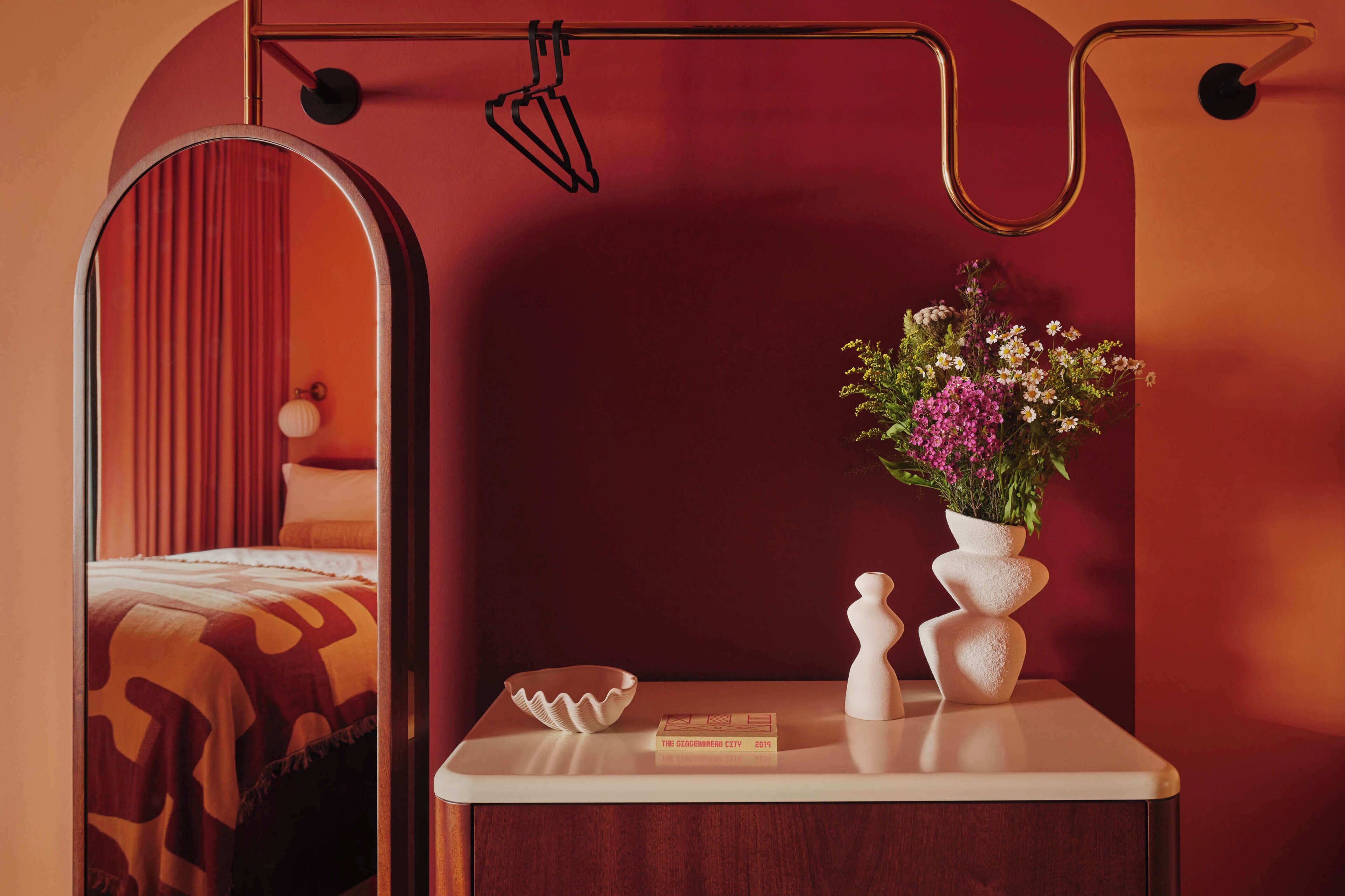 A bedroom with a bed, a bedside table with a book, a mirror, and a vase with flowers.