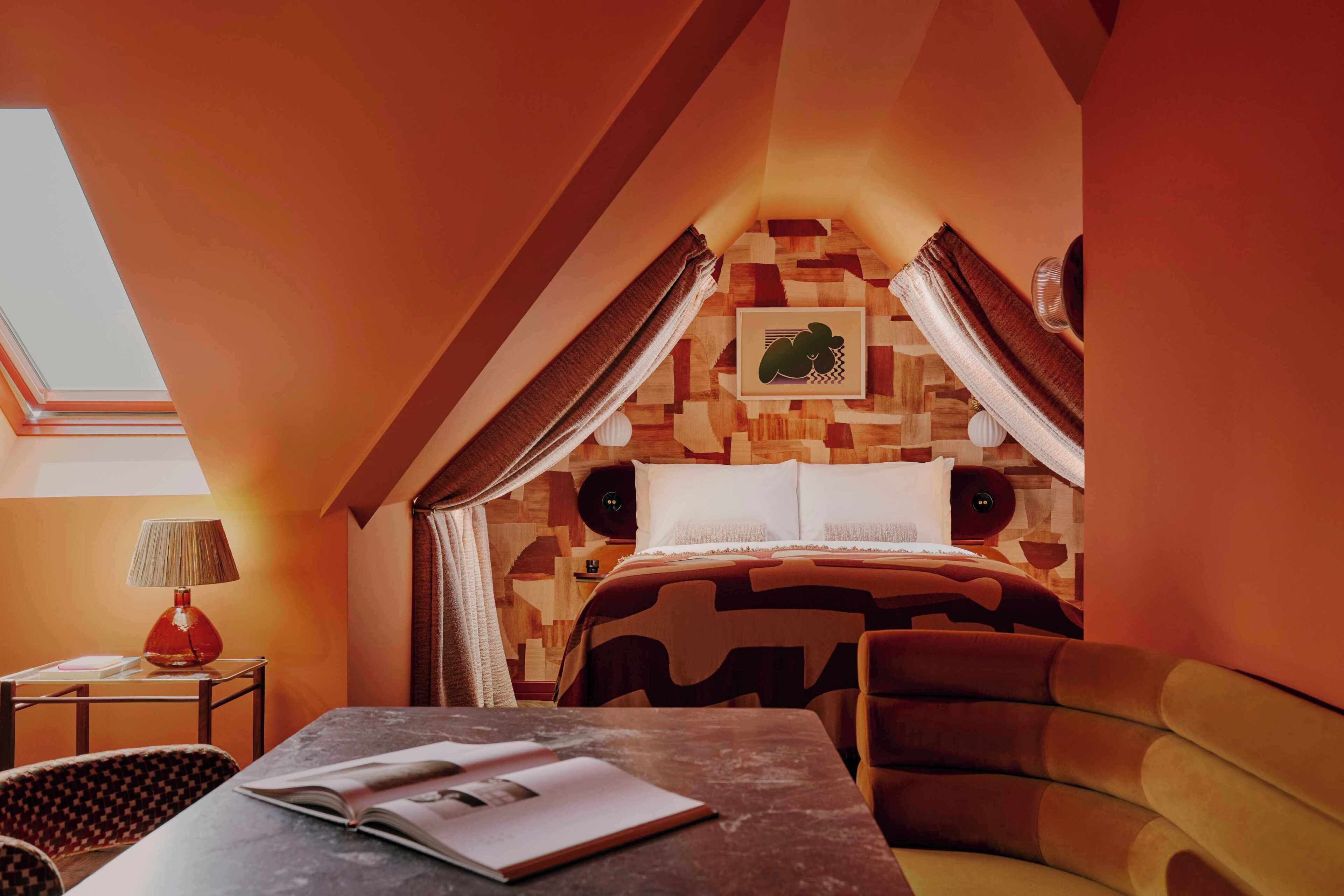 Um quarto acolhedor com teto inclinado, uma cama com cabeceira estampada, uma mesa com um livro, um abajur e uma cadeira.