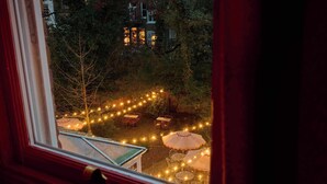 A view through a window of an outdoor seating area with tables and umbrellas, surrounded by trees and a building in the background. - Ember Locke Kensington (London)