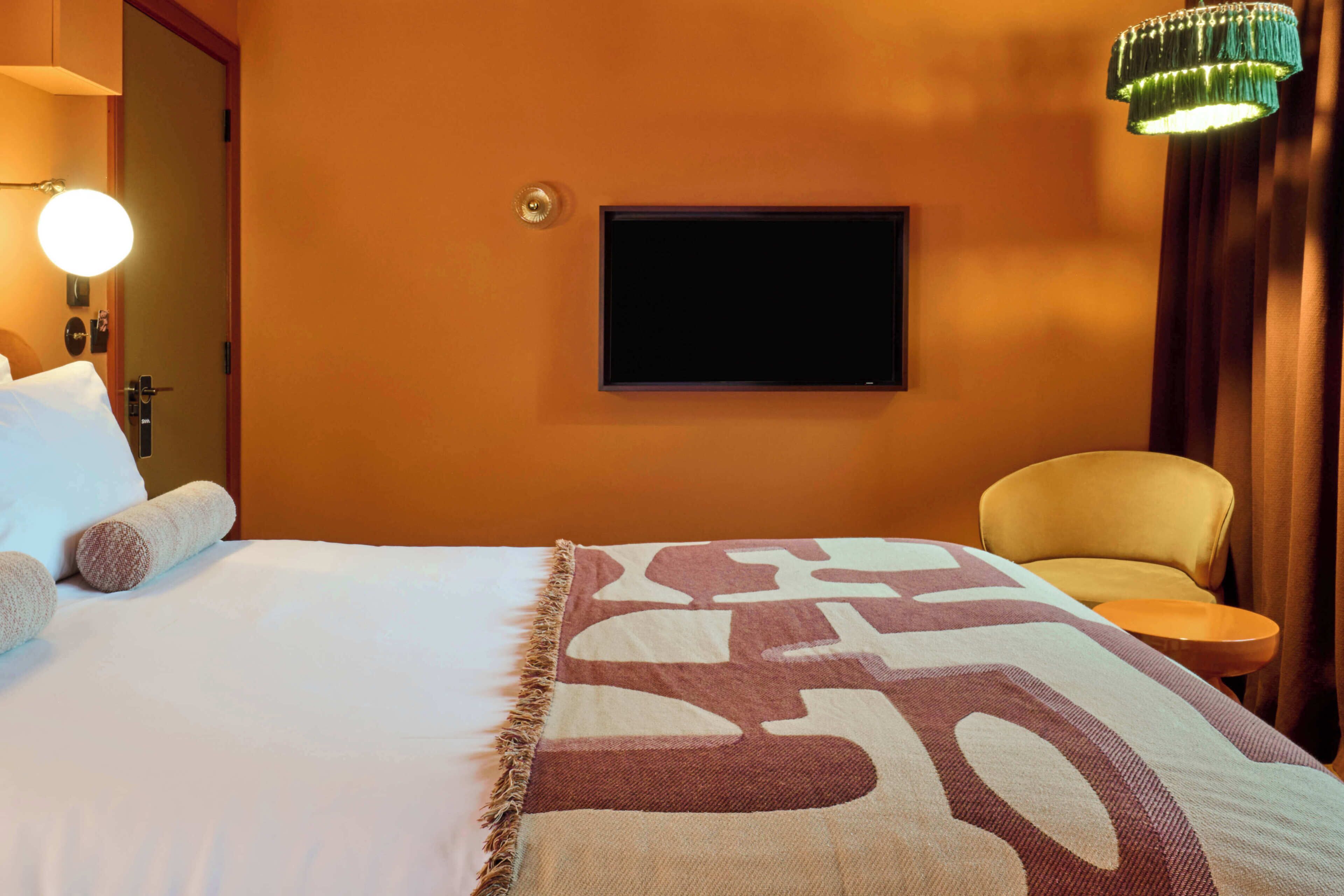 Una habitación de hotel con cama, televisor montado en la pared, una silla y una mesita.