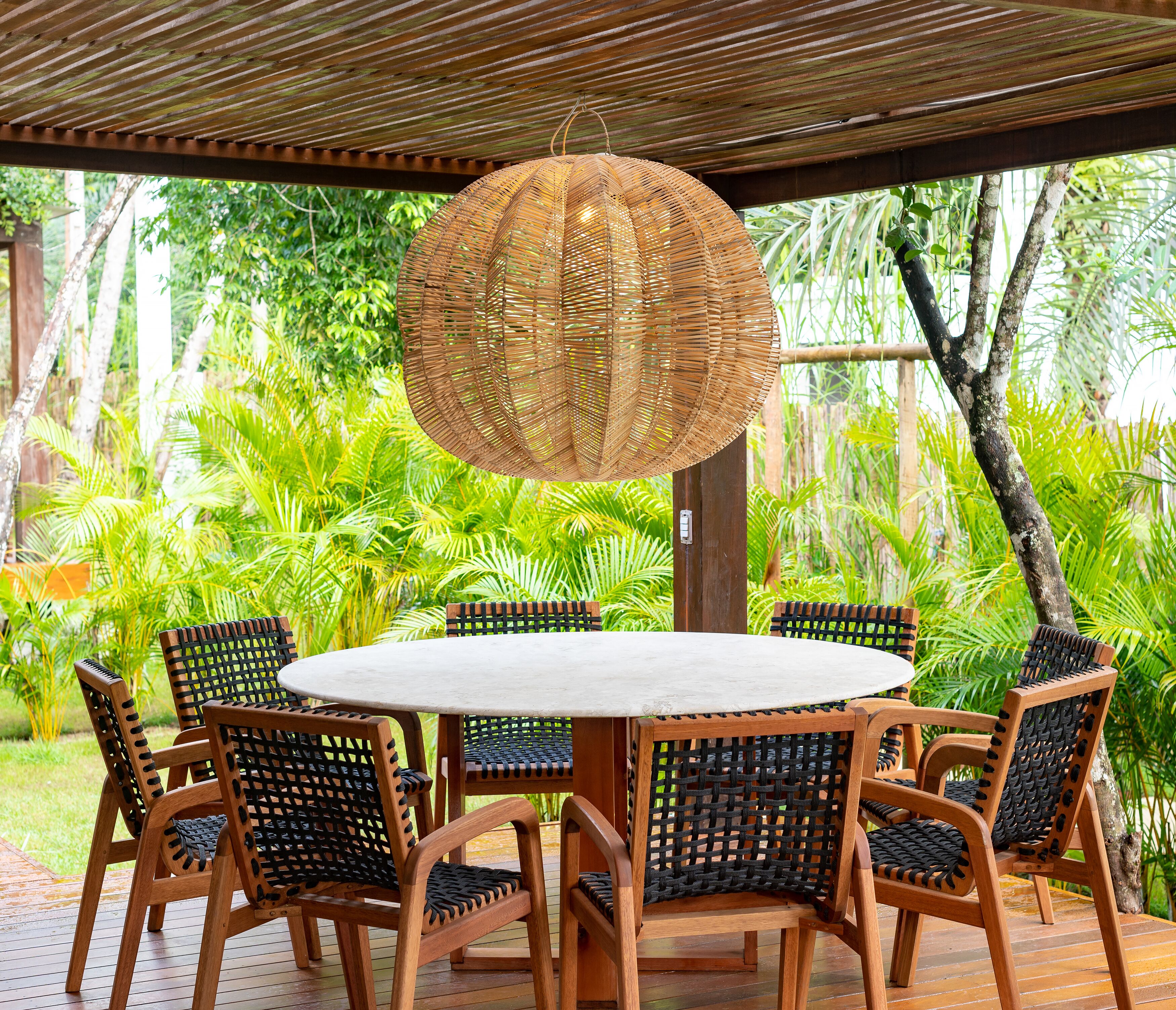 A round wooden table with six chairs, each with a woven backrest, under a wooden pergola with a hanging woven light fixture.