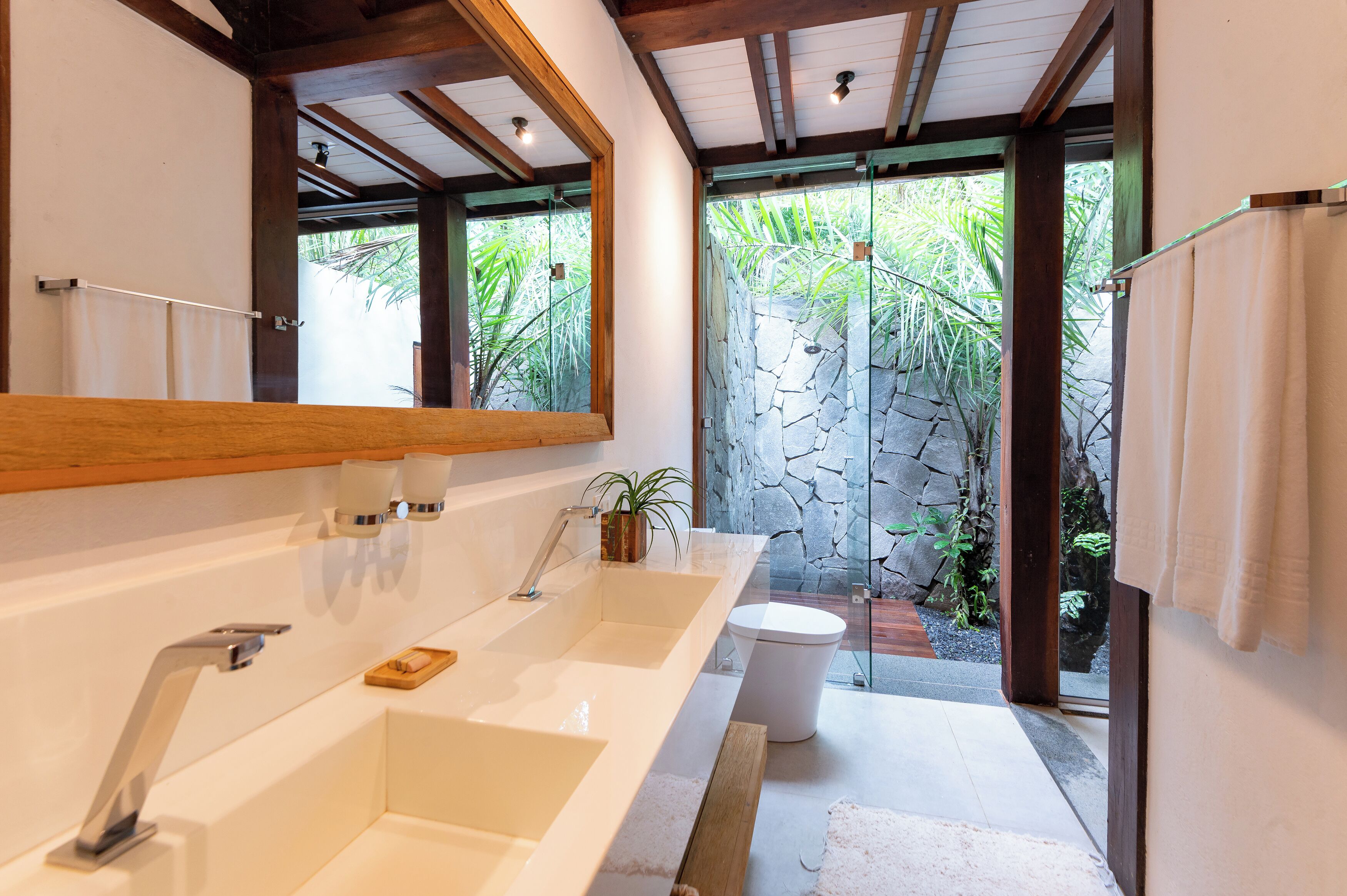A modern bathroom with a large mirror, two sinks, a toilet, and a shower area with a stone wall and tropical plants.