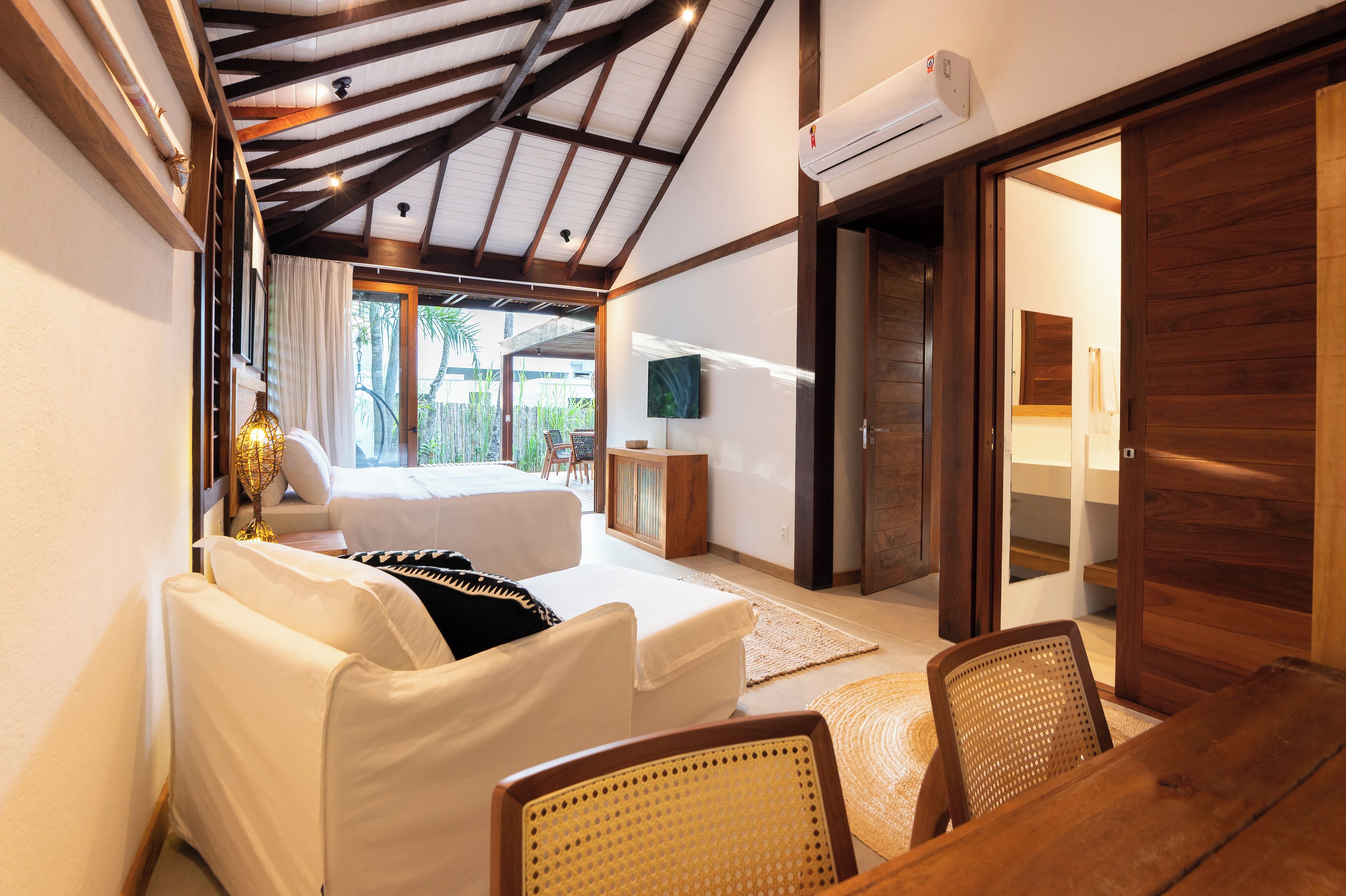 Un dormitorio con cama, una zona de estar con sillas, televisor y puerta corredera que da a un balcón.