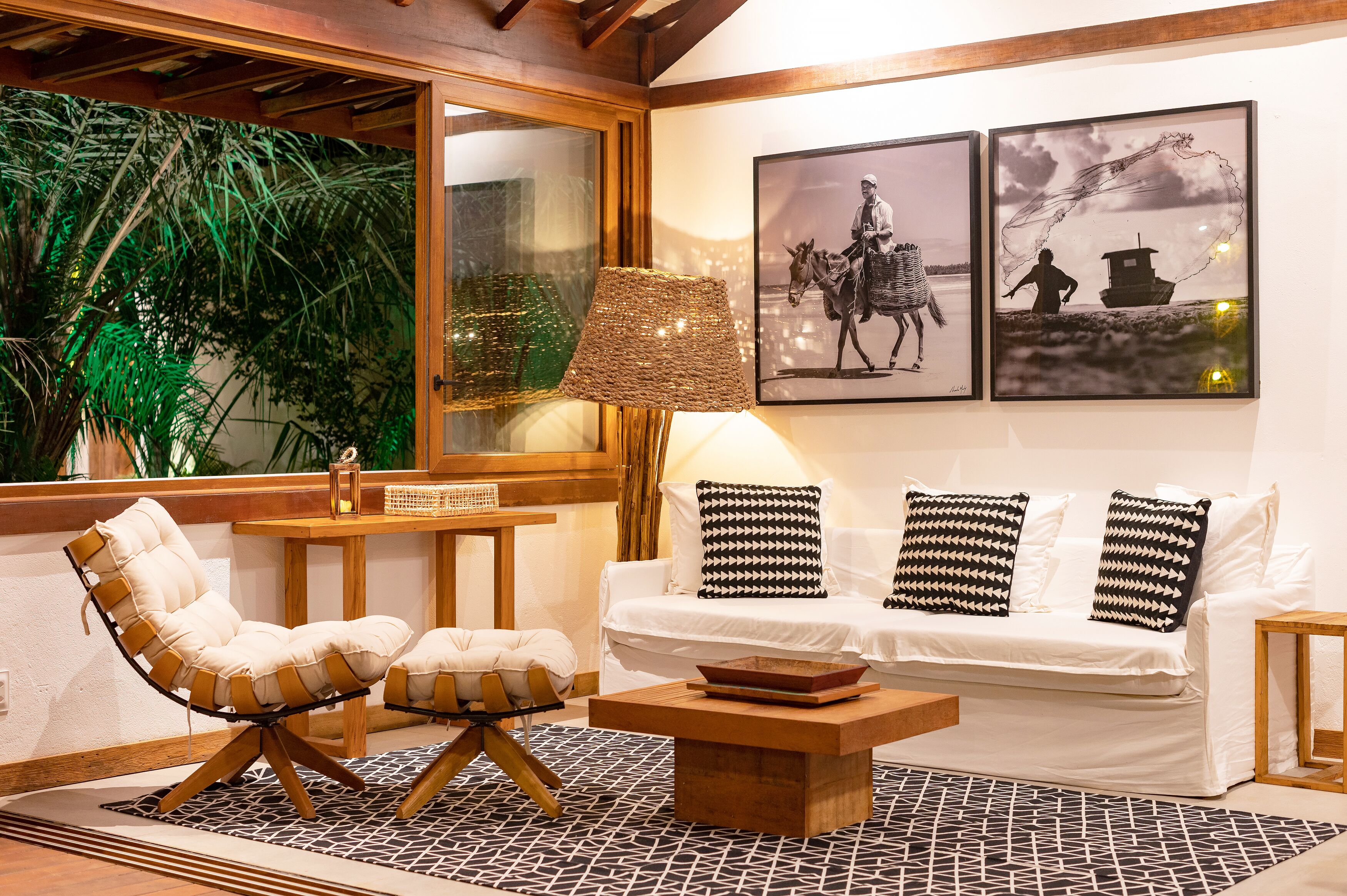 Un salón acogedor con un sofá blanco, un sillón con cojines, una mesa de centro de madera y un ventanal con vistas a follaje verde.