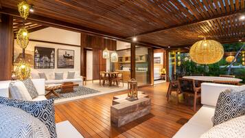 A spacious indoor area with wooden flooring, a seating area with white sofas and patterned cushions, a dining table with chairs, and a large hanging light fixture.