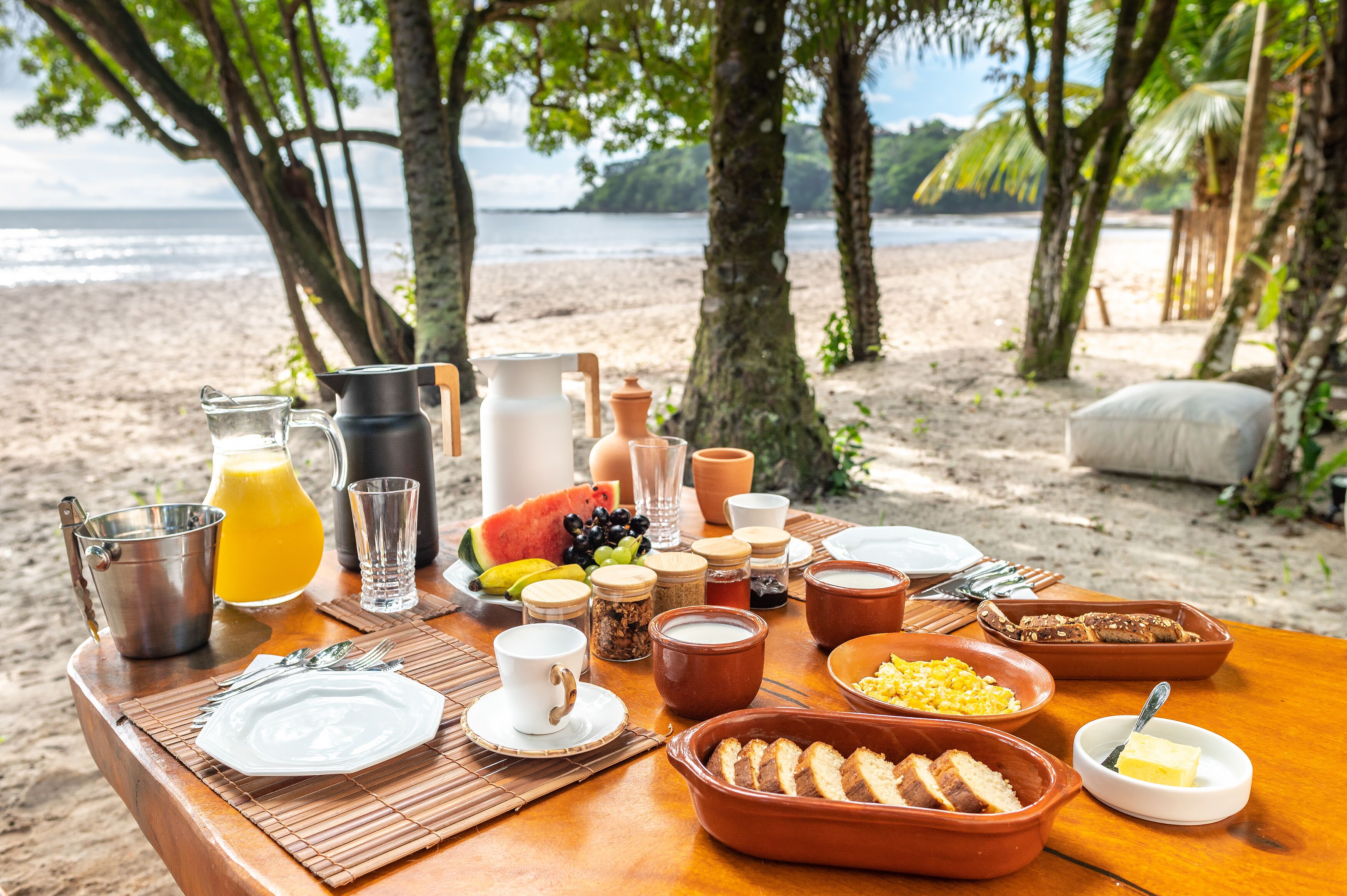A breakfast setup on a wooden table with various food items and drinks, overlooking a beach with palm trees and the ocean.