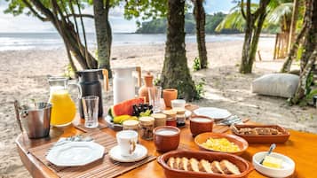 A breakfast setup on a wooden table with various food items and drinks, overlooking a beach with palm trees and the ocean.