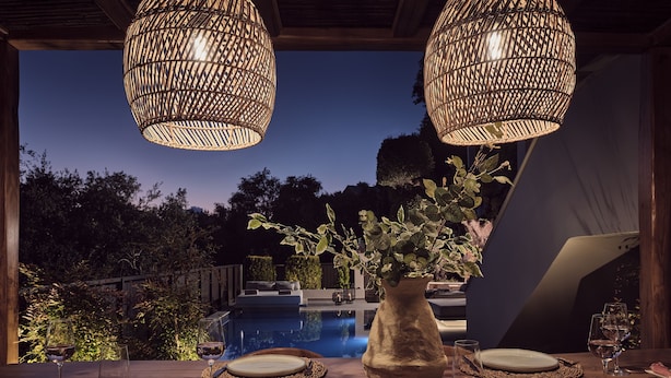 Een terras met een houten tafel gedekt voor twee personen, een vaas met groen en twee rieten hanglampen.