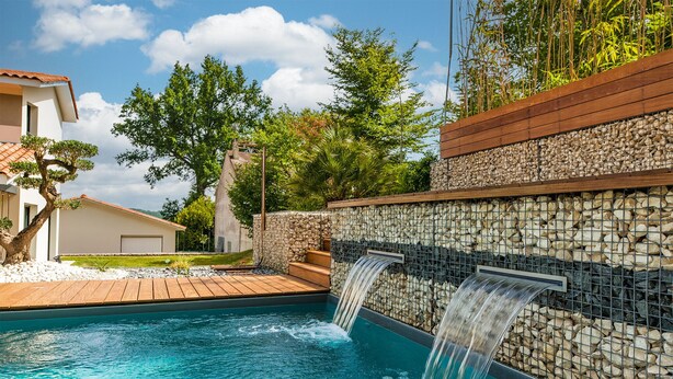 Una piscina con cascada, rodeada por un muro de piedra y una terraza de madera, con una casa al fondo.