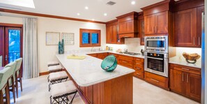 A kitchen with wooden cabinets, a large island, a built-in oven, and a dining area with bar stools. - Casa Luna 17: Luxe Oasis South of 7Mile Beach w/ Secret Beach Cove, Pool + Heated Spa (Grand Cayman)