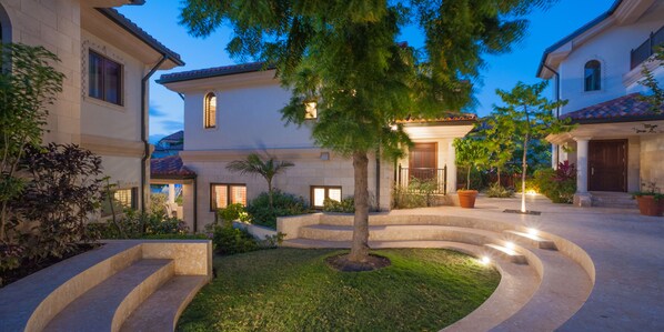 A courtyard with a central tree, surrounded by a stone pathway, and adjacent buildings with multiple windows and doors. - Casa Luna 17: Luxe Oasis South of 7Mile Beach w/ Secret Beach Cove, Pool + Heated Spa (Grand Cayman)