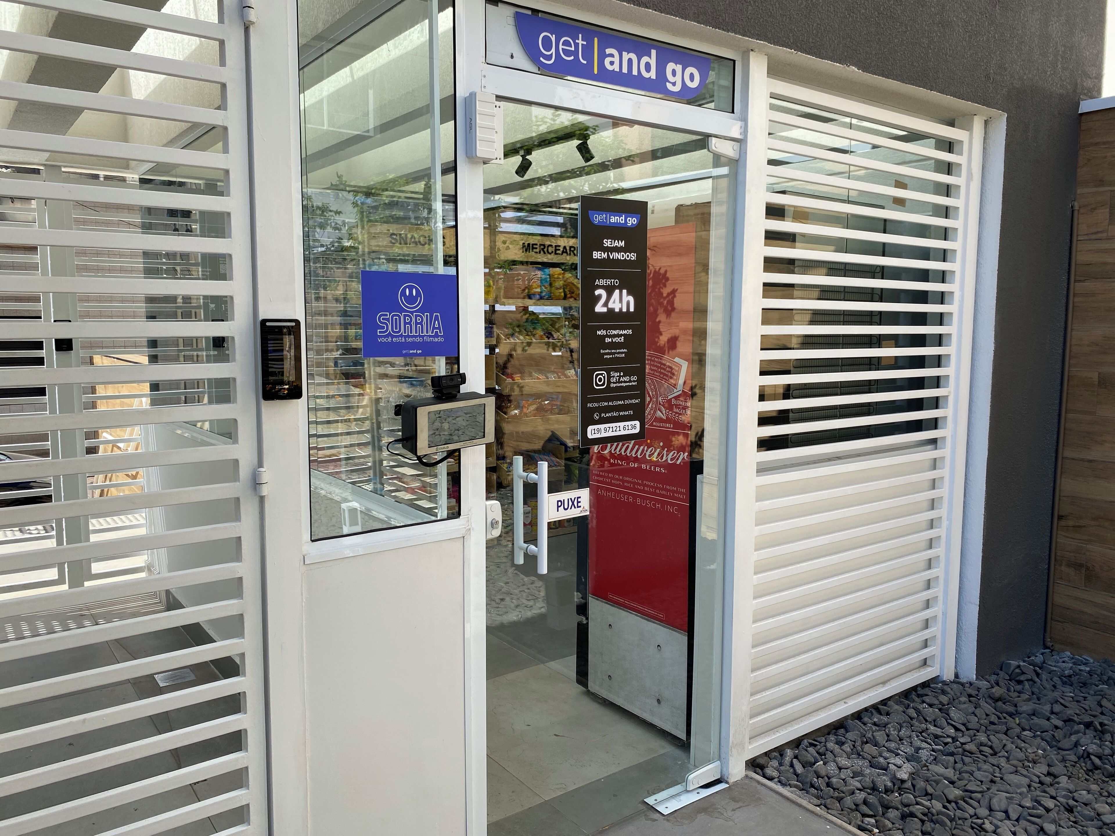 Une devanture de magasin avec une enseigne indiquant « vente à emporter », une caméra de surveillance et une autre indiquant les heures d’ouverture : « 24h/24 ».