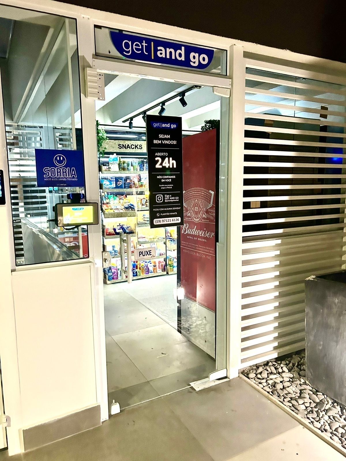 Entrada de una tienda con un cartel que dice 'get and go', un letrero de '24h' indicando que está abierta las 24 horas y un cartel 'Sorria' que muestra una carita sonriente.