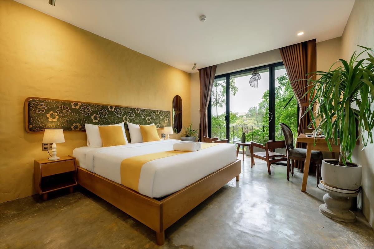 A hotel room with a large bed, a nightstand, a desk, and a potted plant.