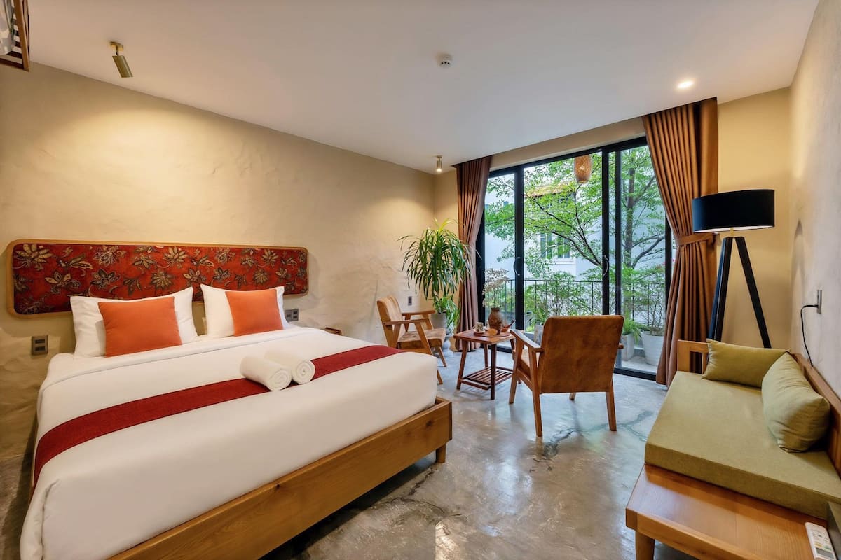 A hotel room with a large bed, a sofa, a small table, and a chair. There is a window with curtains and a view of trees outside.