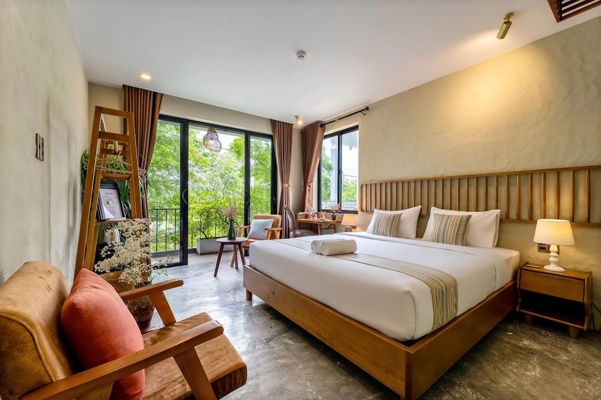 A modern hotel room with a large bed, a wooden headboard, a desk, and a view of greenery outside.
