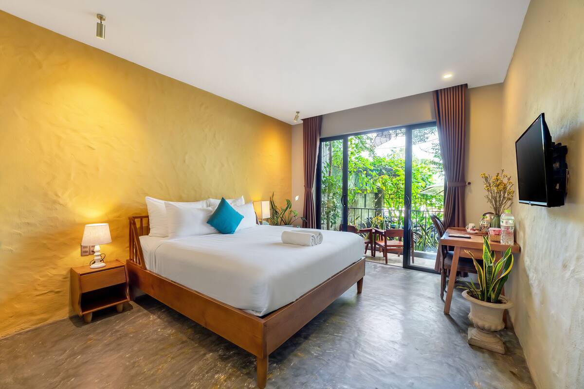A hotel room with a large bed, a desk, a chair, a TV, and a potted plant.