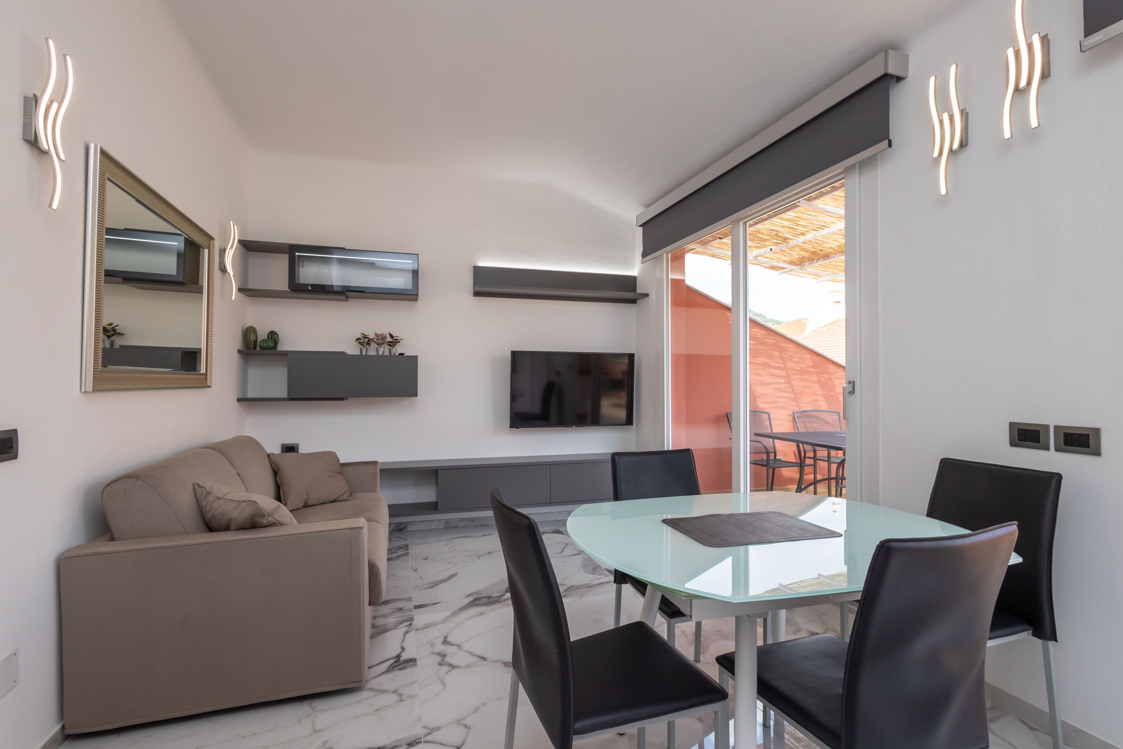 A modern living room with a glass dining table, black chairs, a beige sofa, a flat-screen TV, and a large mirror.