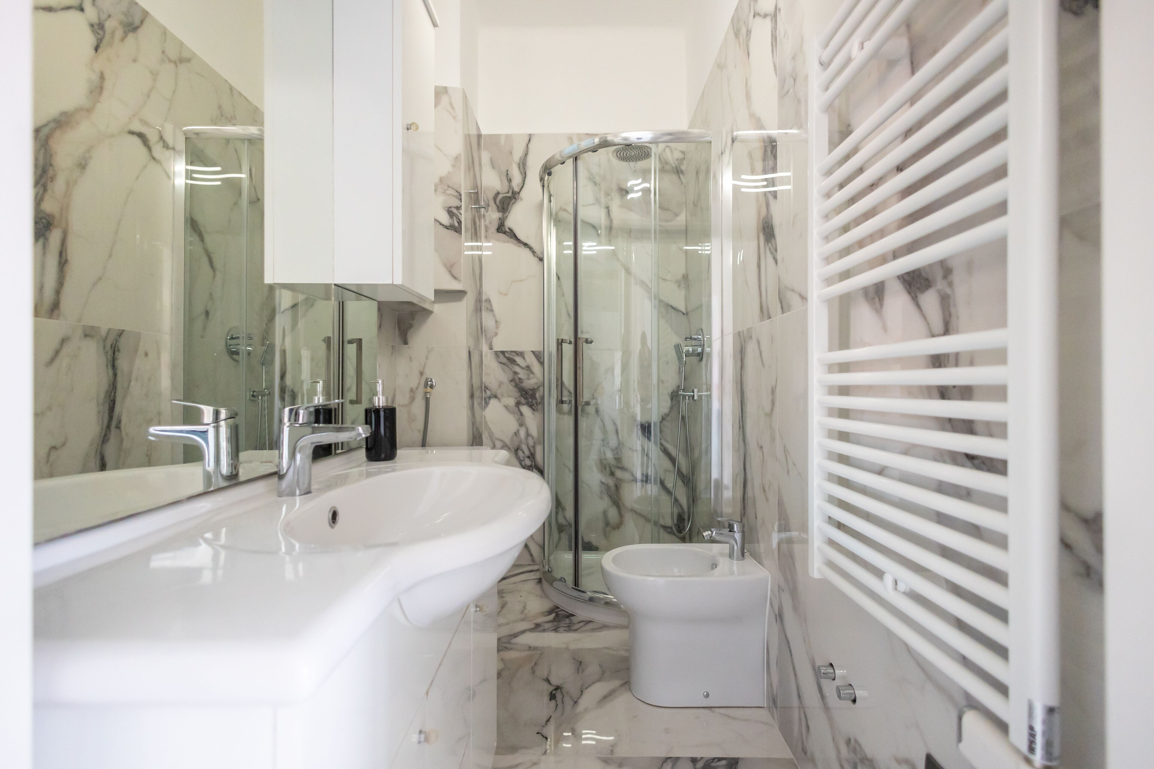 Un bagno moderno con rivestimento in marmo, cabina doccia in vetro, bidet e lavandino bianco.