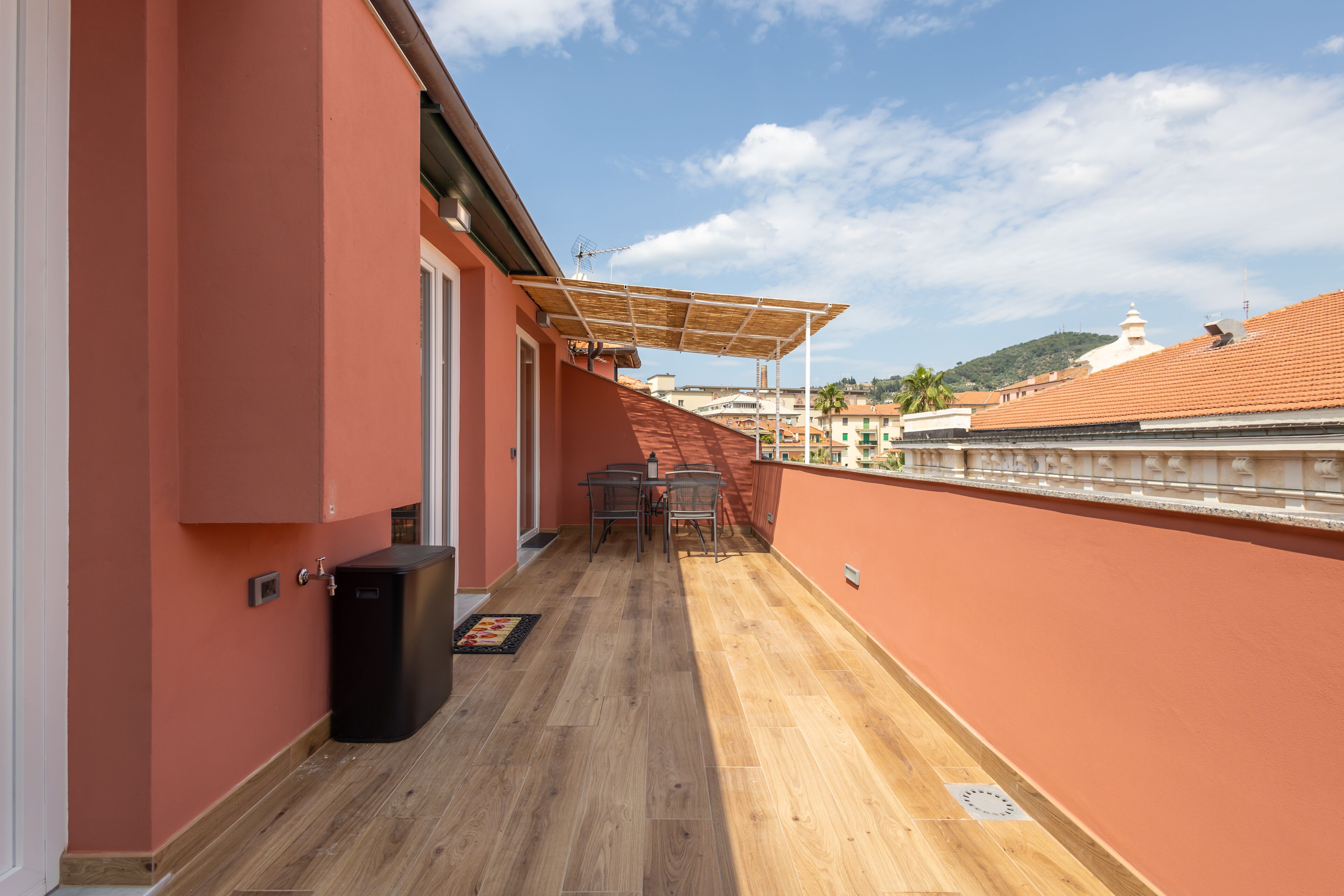 A balcony with a wooden floor, a table and chairs, and a black trash bin.
