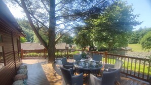 A wooden deck with wicker furniture, a round table, and a stone wall in the background. - Fellhaven  -  a holiday lodge that sleeps 6 guests  in 3 bedrooms (Windermere)