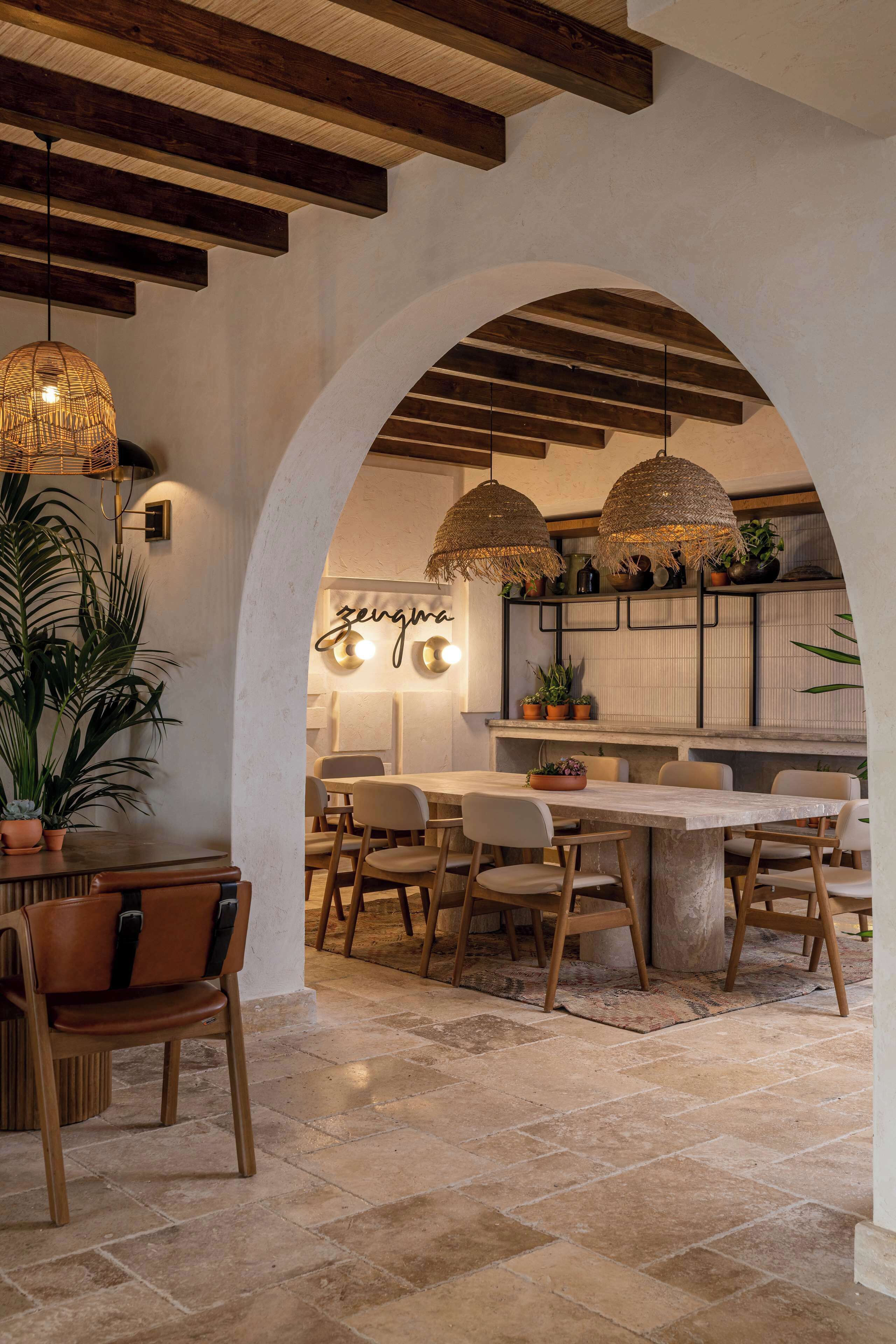 A dining area with a large table, chairs, hanging wicker lamps, and potted plants.
