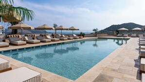 Uma área à beira da piscina com espreguiçadeiras e guarda-sóis, piscina cristalina e vista para o oceano e colinas ao fundo.